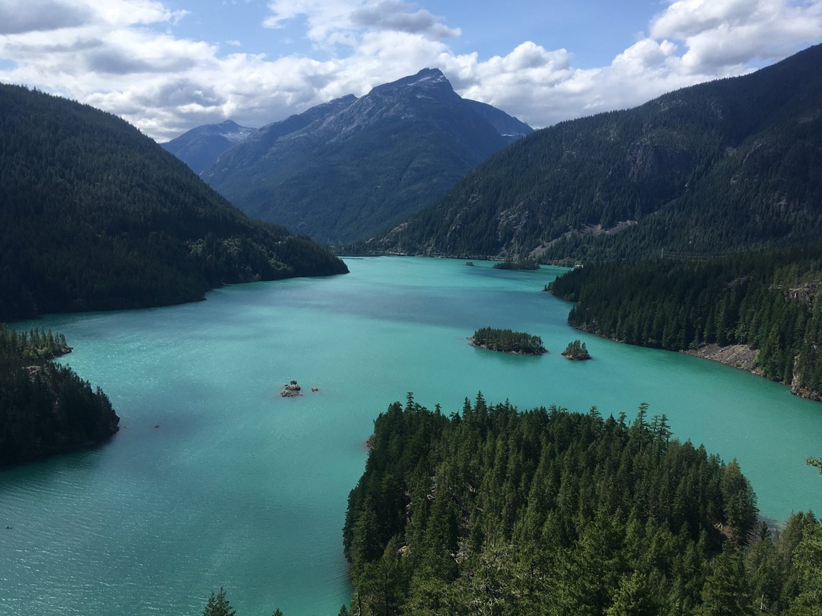 A turquoise glacial lake with small forested islands, surrounded by dark forested mountains under a partly cloudy sky