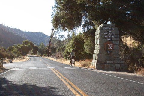 Visitor seen along a paved roadway riding a bicycle past a park service sign made into a rock pillar