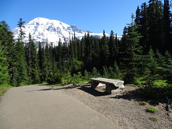 A wide paved trail with a low log bench to the right. Mount Rainier rises above the trail.