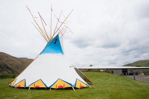 Color photograph of a tipi pitched on the grass in front of a low-slung building.