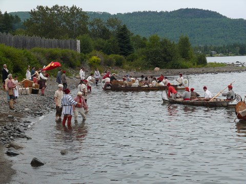 Canoes and people wearing historic clothing at the edge of a lake, next to a stockade.