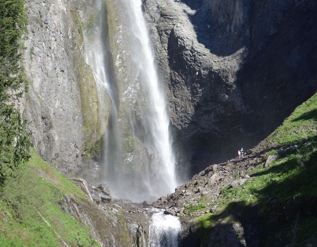 A white plume of water descends into a rocky canyon with hikers viewing it from a trail.