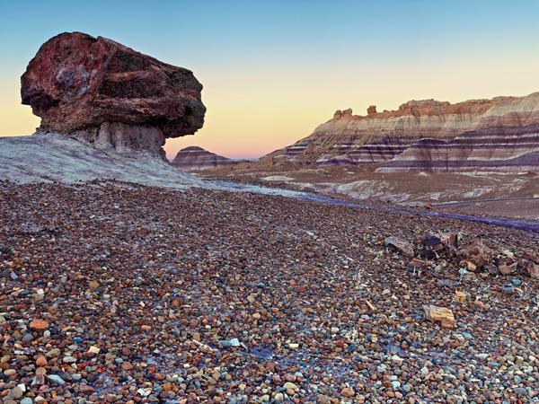 A pedestal log balances in front of a blue, purple, and gray banded badland at twilight.