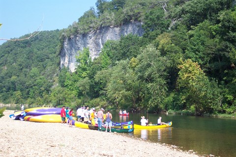 pile of canoes on gravel riverbank with trees and cliff in background