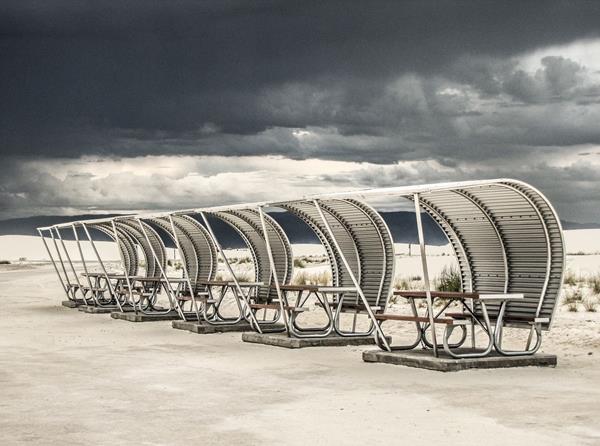 a row of curved metal structures provides shade to picnic tables on a concrete pad.
