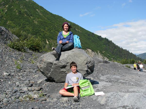 Two kids sitting down. One is on a boulder and the other on the ground.