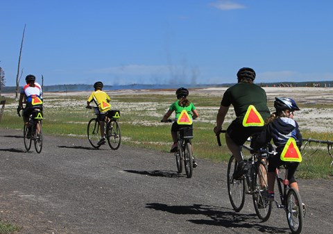 Five cyclists ride on a trail with steam rising from a geysers in the background.