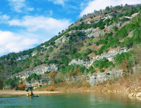 Two paddlers in a kayak passing by a rocky and vegetated bluff.