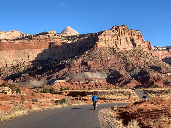 Person on bicycle on paved road with cars approaching and colorful cliffs above, and blue sky.
