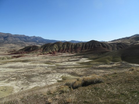 Red and green striped hills with shrubs in the foreground and clear blue skies in the background.