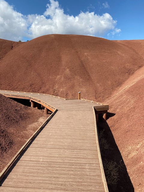 A wooden boardwalk goes between two red claystone hills. Cloudy blue skies are in the background.