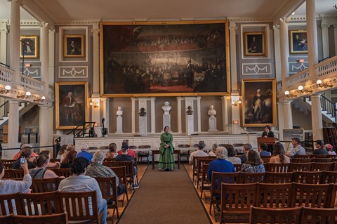 A woman in a green 1800s dress stands in front of the stage and paintings in the Great Hall