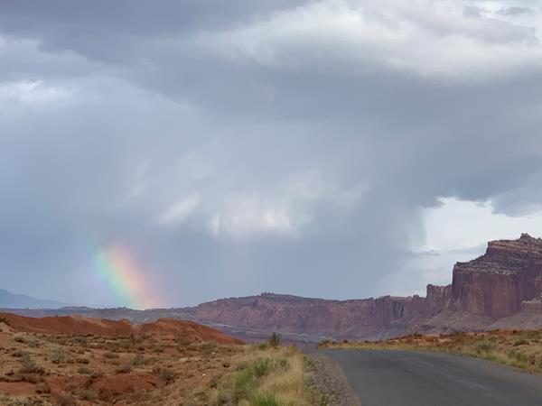 Stormy skies with a small spot of a rainbow, red cliffs, and a blacktop road.