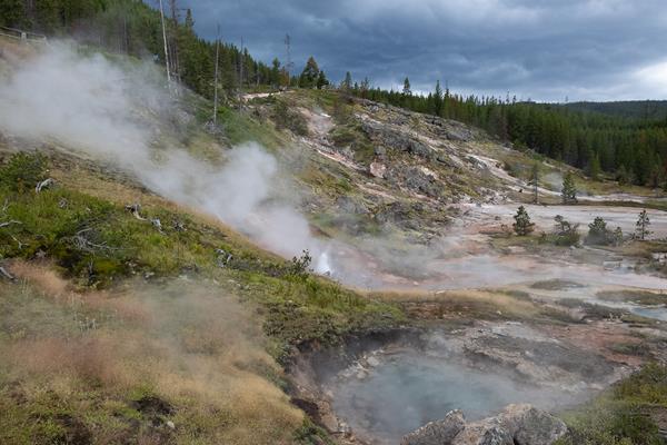 Hot springs produce steam at the base of a hill under a stormy sky.