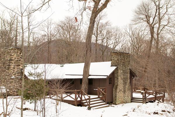 An historic cabin with a large porch sits in the middle of a snow-covered forest.