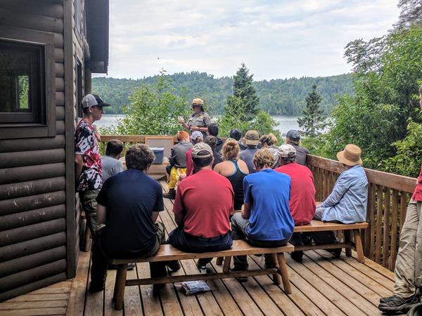 A park ranger gives a program to a group of people on a porch with Washington Harbor behind.