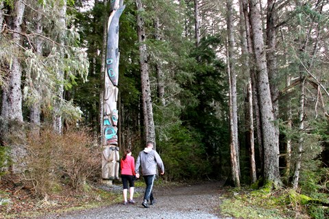 A man and woman walk by trees and a totem pole on a gravel path