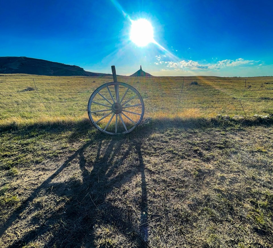 A wagon wheel rests on barbed wire fence with Chimney Rock and blazing sun in the background.