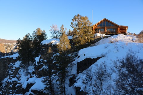 A log structure with large windows sits on the edge of a steep, snowy canyon wall.