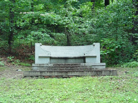 A granite bench sits atop a slight rise surrounded by green trees