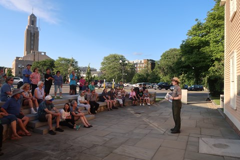 Ranger addressing large group of people sitting on stone steps in front of yellow building
