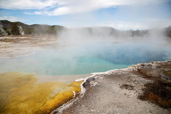 Steam rises off a turquoise blue hot spring with orange microbial mats along the edge.