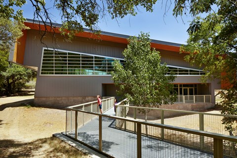 People stand on an elevated walkway leading to a large building with many windows and slanted roof.