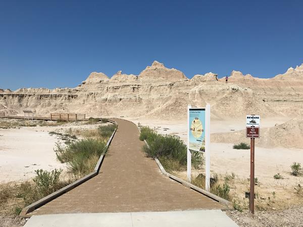 A boardwalk extends towards badlands formations under blue sky amid exhibit and rattlesnake sign.