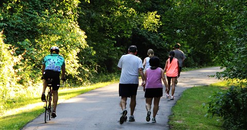 5 people walking away from viewer on the right side of the trail. 1 bicyclist passes the 5 walkers.