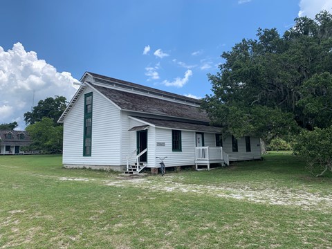 A white building with pitched roof in an open field with a large live oak to the right.