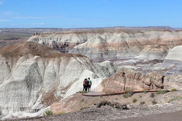 Hikers walk amidst gray, blue, and purple badlands along a trail under a blue sky.