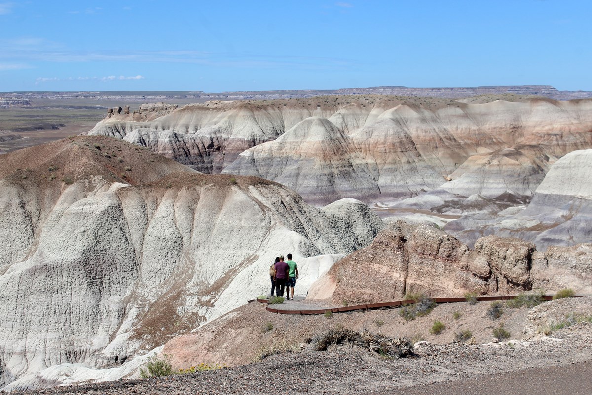 Hikers on a trail among gray, blue and purple badlands under a blue sky.