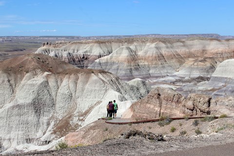 Hikers on a trail among gray, blue and purple badlands under a blue sky.