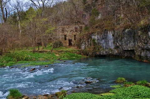 a rock building in distance with blue spring flowing in front, green vegetation around spring