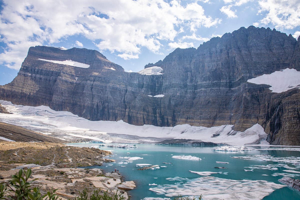 A turquoise lake with floating pieces of ice sits at the base of snow-speckled, striated cliffs.