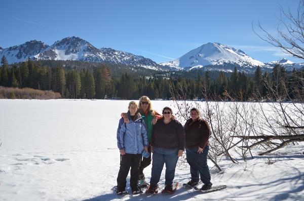 Four women in snowshoes standing on the edge of a snow-covered lake backed by volcanic peaks.
