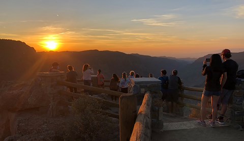 Visitors at a log-and-stone railing watching the sun set over the canyon
