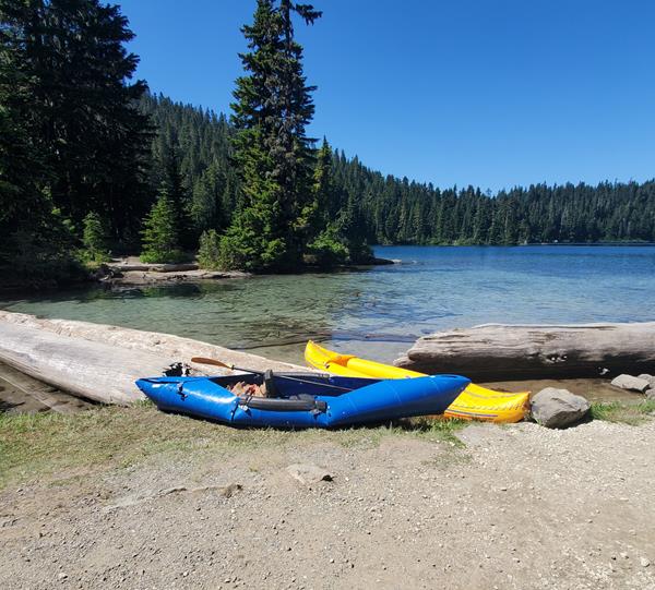 A ten-foot long blue kayak sits on the bank of a lake in front of an inflatable yellow kayak.