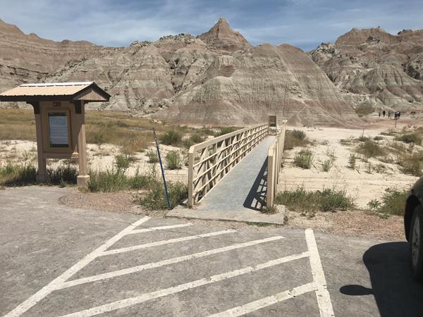 Information board sits alongside boardwalk which extends into badlands formations under blue sky.
