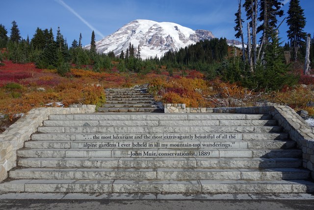 Stone steps lead toward the glaciated peak of Mount Rainier framed by red-orange fall foliage.