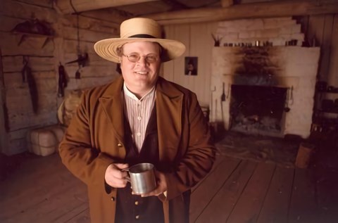Man in straw hat brown wool coat, vest and striped shirt holds a tin cup