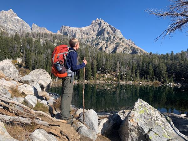 Backpacker standing at the edge of an alpine lake with mountains and trees surrounding