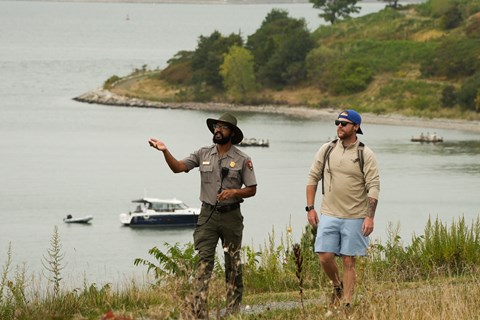 A uniformed ranger walks with a guest on a grassy path on and island, boats in the water behind them