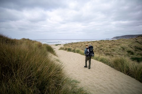 A photograph of a man carries a young child on his back while walking along a path over a sand dune.