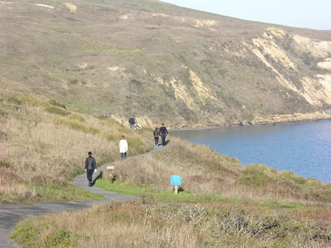 A photograph of five people walking along a narrow blufftop trail with a bay on the right.