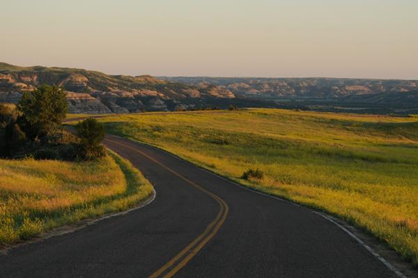 A road curves out of sight around some buttes. Yellowish grass grows on both sides of the road.
