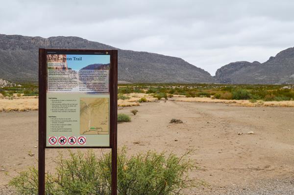 A metal sign with trail information is in front of a flat desert trail that leads to a canyon.