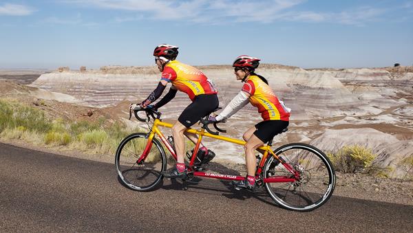 Two people on a yellow and red tandem bike ride past blue striped badlands.