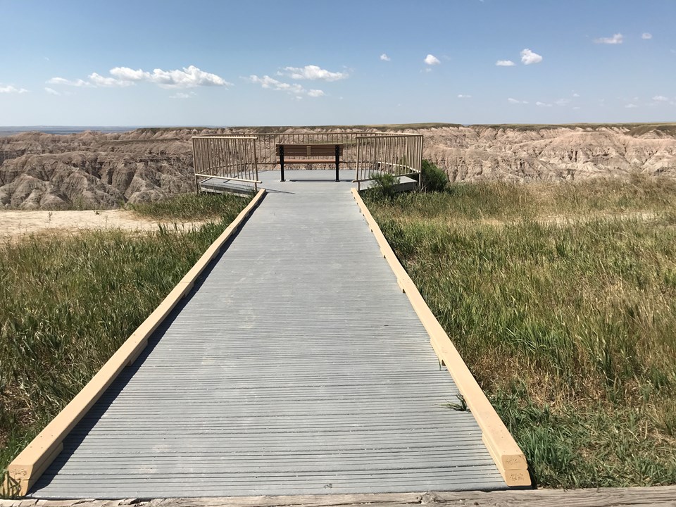 A single boardwalk extends out to a bench which sits against a vast landscape under blue sky.