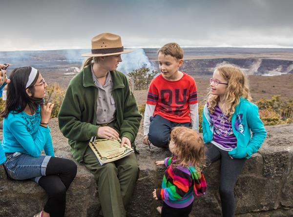 A park ranger sits on a stone wall on the edge of a volcanic caldera surrounded by children